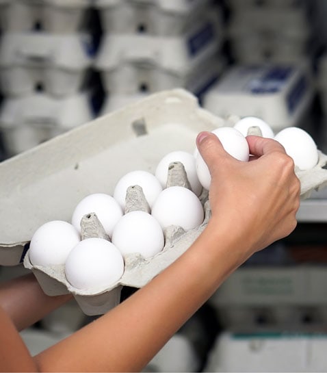 A person holding an open carton of white eggs with stacks of egg cartons in the background.